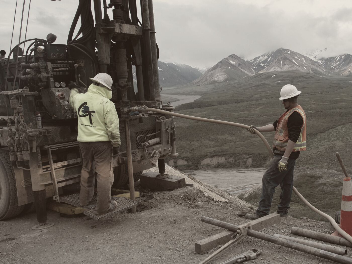 Two drillers working at a track-mounted core rig on a mountain road, with drill rods and a hose on the ground and a river valley behind
